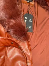 Tapado de cuero sintético color bordó, con cuello y puños de piel sintética desmontables. Presenta cierre frontal con botones y cinturón ajustable con hebilla forrada en el mismo material.