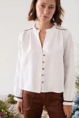 Blusa blanca de lino con escote en V y cuello con volados. Mangas largas con puños elastizados adornados con volados y detalles de ribetes en tono marrón oscuro. Cuenta con cierre frontal de botones.
