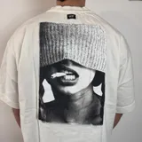 Remera negra de algodón pesado, corte oversize, con un gran estampado en la espalda en blanco y negro que muestra la parte inferior del rostro de una persona con un gorro tejido cubriendo los ojos y un cigarrillo en la boca.