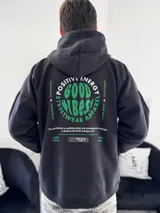 Canguro oversize negro con capucha y bolsillo tipo canguro frontal. Presenta un gran estampado en la espalda con la leyenda "Good Vibes Positive Energy Streetwear Apparel" en tonos verdes y blancos.
