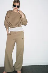 Pantalón de felpa color beige, tiro medio, con cintura elástica, bolsillos laterales ocultos y pierna ancha. Presenta un parche pequeño con un logo en la parte delantera.