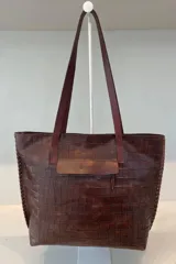 Bolso tipo tote de cuero vacuno con textura de reptil, color marrón. Presenta detalles de pasamanería en los bordes laterales y una borla de flecos de cuero colgante. Cuenta con cierre superior.