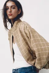 Chaqueta bomber de color beige con diseño a cuadros blancos, cuello subido, manga larga con trabillas en los hombros, bolsillos delanteros y bajo con elástico. Cierre frontal con cremallera y botones a presión.