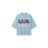 Remera de algodón celeste con corte oversize, cuello redondo y mangas cortas amplias. Presenta un estampado gráfico frontal con la inscripción "USA" en azul y rojo, acompañada de la palabra "ESSENTIALS" en tono beige.
