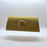 Bolso tipo clutch rígido y alargado color mostaza, con solapa y un aplique circular central con piedra de resina sobre una base trenzada y dorada. Incluye asa de cadena desmontable.