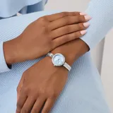 Reloj analógico de estilo joya con caja redonda y malla de acero inoxidable plateado. La esfera es de color celeste con textura sutil, números romanos en las posiciones 6 y 12, y detalles de cristales engastados en el bisel y en los eslabones con forma de corazón.