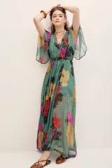 Vestido largo verde con estampado floral multicolor, escote en V cruzado, mangas amplias tipo capa y detalles metalizados.