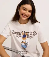 Remera blanca de algodón con cuello redondo y manga corta. Presenta un estampado central con texto que dice "Sunny Mornings Slow Days" y debajo un dibujo de un conejo con gorra azul, shorts y una mochila.