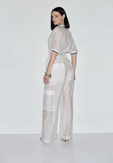 Mono largo de mujer confeccionado en organza transparente, con escote en V con cierre frontal, mangas cortas con puños acanalados a rayas y cintura ajustable con cordón. Presenta un diseño de pierna ancha con bolsillos tipo cargo en los laterales.