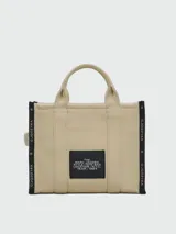 Bolso tipo tote bag de Marc Jacobs en color beige, confeccionado en lona resistente. Presenta el texto "THE TOTE BAG" y "MARC JACOBS" impreso en la parte frontal en color negro. Cuenta con asas superiores y una correa de hombro ajustable y extraíble, además de ribetes laterales negros con inscripciones.