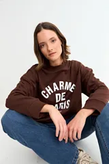 Buzo de cuello redondo y manga larga, color marrón chocolate, con estampa frontal en color blanco que dice 'CHARME DE PARIS'.