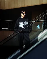 Hoodie negro de corte amplio con capucha y bolsillo frontal tipo canguro. Presenta un estampado gráfico artístico en blanco y negro en el pecho y un pequeño detalle de etiqueta naranja en la parte inferior.