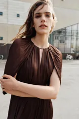 Vestido corto de tejido satinado color marrón, con cuello redondo, mangas largas con diseño fluido y cintura elástica. Presenta una abertura frontal y cierre posterior con botón.