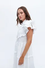 Vestido midi de lino blanco con diseño de broderie, cuello alto, cierre frontal con botones y volados decorativos en los hombros y el cuerpo.