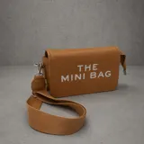Cartera tipo bandolera de cuero sintético texturizado color tostado, con la inscripción 'THE MINI BAG' estampada en blanco. Incluye correa de cinta regulable y extraíble, y herrajes metálicos plateados.