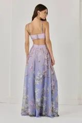 Vestido largo de gasa con estampado floral en tonos lila y rosa, con una flor bordada en 3D en el busto, corte en la cintura y falda amplia con abertura lateral. Cuenta con botones y cierre trasero con cremallera invisible.