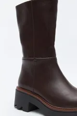 Bota de caña media en cuero sintético color negro, con diseño minimalista y suela robusta dentada con detalle de borde en color marrón.