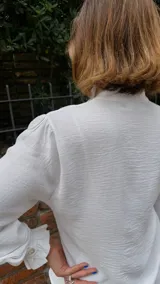 Camisa blanca de textura sutil, con cuello alto tipo victoriano y detalle de volados asimétricos a lo largo de la botonadura frontal. Presenta mangas largas con puños terminados en volados.