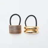 Set de dos colitas elásticas para el pelo. Una presenta un aplique rectangular con acabado marmolado en tonos marrones y bordes dorados, mientras que la otra cuenta con un aplique metálico dorado de diseño doble redondeado.