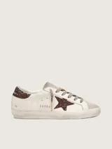 Zapatillas Golden Goose modelo Super-Star, color blanco con detalles en marrón con glitter. Confeccionadas en cuero vacuno, con estrella lateral y contrafuerte de talón brillantes, punta y lengüeta de gamuza, y efecto envejecido en la suela.