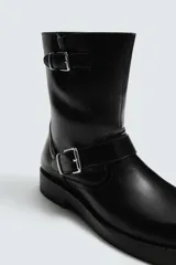 Bota de media caña en color negro, con diseño de horma cuadrada y suela con ligero tacón. Presenta dos cintas ajustables con hebillas metálicas, una en la parte superior de la caña y otra en la zona del empeine.