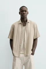 Camisa de calce relajado confeccionada en tejido con mezcla de algodón y lino, con diseño de rayas verticales en tonos beige. Presenta cuello bowling, manga corta, aberturas laterales en el bajo y cierre frontal con botones.