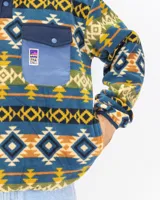 Buzo polar de cuello alto con cierre frontal de botones a presión, confeccionado en fleece sintético. Presenta un estampado geométrico multicolor con predominancia de tonos azules, amarillos y verdes. Incluye un bolsillo frontal tipo cargo con solapa y cierre de botón, además de bolsillos laterales ocultos.