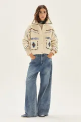 Campera de lana merino hervida en color crudo, con diseño de cuello camisero, cierre frontal de botones y bolsillos frontales. Presenta bordados artesanales con motivos geométricos en tonos marrón y azul en el frente, espalda y mangas.