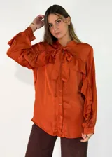 Camisa de satén color naranja óxido, con cuello mao y lazo para anudar. Presenta volados decorativos en el frente y en las mangas, las cuales son largas y terminan en puños abotonados.