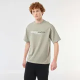 Remera de algodón verde militar, de corte holgado y manga corta, con cuello redondo y estampado frontal con texto en blanco y negro.