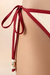 Calzón de bikini color blanco marfil, con ribete rojo oscuro y tiras laterales con detalle de piedras doradas.