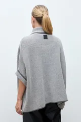 Capa de tejido de punto en color gris, con diseño abierto, cuello alto y cierre frontal mediante una hebilla metálica. Presenta bordes con ribete en color negro.
