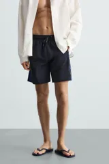 Bañador largo negro con cintura elástica ajustable mediante cordón. Presenta un panel lateral con estampado de cuadros en tonos azul y negro. Incluye bolsillos frontales y un bolsillo trasero con solapa.