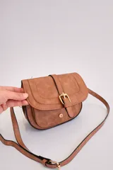 Bandolera de tamaño pequeño en color camel con acabado texturizado. Presenta un diseño de solapa frontal con detalle de hebilla decorativa y cierre magnético. Incluye una correa larga ajustable y herrajes metálicos en tono dorado.