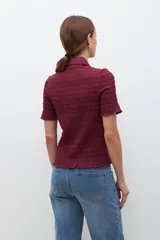 Camisa de manga corta en color bordó, confeccionada en tejido con textura de punto smock. Presenta cuello clásico, cierre frontal con botones y un ajuste entallado al cuerpo.