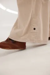 Pantalón sastrero de tiro medio confeccionado en pana de algodón. Presenta un diseño de pierna ancha con costuras verticales decorativas, bolsillos delanteros y traseros, y un detalle de aplique con ojalillo metálico en el ruedo de una de las piernas.