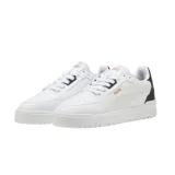 Championes urbanos Puma modelo Shuffle Downtown, de diseño clásico y perfil bajo. Presentan una capellada blanca con detalles en negro en la zona del talón y el logo de la marca en dorado. Cuentan con suela de goma y sistema de cordones.