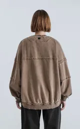 Buzo de cuello redondo color marrón claro, confeccionado en algodón premium con textura acanalada. Presenta costuras expuestas y detalles deshilachados en cuello, puños y ruedo, con un calce oversize y mangas abullonadas.