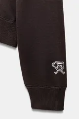 Sudadera de cuello redondo y manga larga con hombro caído, confeccionada en mezcla de algodón. Presenta un diseño liso en color marrón oscuro con un pequeño estampado gráfico a contraste en el pecho y detalle en el puño.