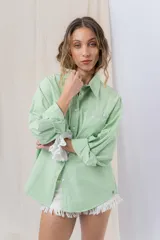Camisa verde pastel de algodón, corte oversize, con cuello camisero, cierre con botones, mangas largas con puños con volados de encaje blanco, dos bolsillos frontales con solapa y ruedo irregular. 