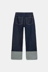 Jeans de tiro bajo y pernera ancha, confeccionados en denim azul oscuro con pespuntes en contraste. Cuentan con un diseño de cinco bolsillos, cierre frontal con cremallera y botón metálico, y destacan por su amplio doblando en el bajo que deja ver el revés de la tela con detalles de costura selvedge.