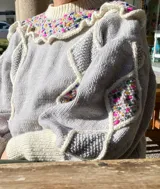 Sweater de lana merino gris con detalles bordados a mano de colores en el canesú y en las mangas. Presenta cuello redondo, volados decorativos y diseño de rombos con efecto 3D.