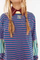 Remera de manga larga con corte boxy, confeccionada en algodón. Presenta un diseño de rayas horizontales en tonos azul y fucsia, con detalles de rayas verticales en los codos, tajos laterales y logo bordado en el pecho.
