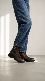 Bota urbana de caña media, confeccionada en una combinación de cuero gastado y gamuza. Presenta una solapa frontal en gamuza, cierre lateral y una tira decorativa con hebilla metálica en el tobillo. Cuenta con una suela de goma robusta y dentada.