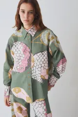 Camisa de corte holgado con cuello clásico y cierre frontal de botones. Presenta un estampado temático de elementos deportivos en tonos verde, rosa y beige, con mangas largas y puños abotonados.