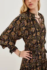 Vestido midi de corte fluido con estampado paisley en tonos oscuros, ocres y rosados. Presenta cuello redondo con lazo de ajuste, mangas largas abullonadas con puños elásticos y falda con volados en el ruedo.