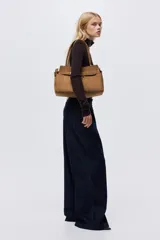 Bolso tote de acabado suave, color marrón, con asas tubulares unidas por una trabilla con hebilla metálica. Presenta una correa decorativa frontal con hebilla metálica y compartimento interior con cierre.