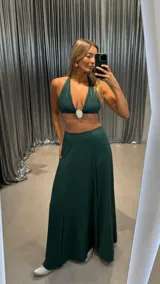 Conjunto de dos piezas color verde oscuro, compuesto por un top tipo bralette con cuello halter y un dije ovalado blanco en el centro, y una falda larga y fluida.