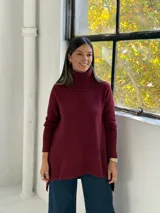 Sweater de punto color bordó con cuello alto tipo polera, diseño de corte evasé con costura central vertical y bajo asimétrico más largo en la parte posterior.