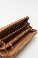 Billetera de formato rectangular con cierre perimetral, confeccionada en material símil cuero de textura suave. Presenta un aplique metálico circular con logo grabado en el frente y tirador de cierre en el mismo material.